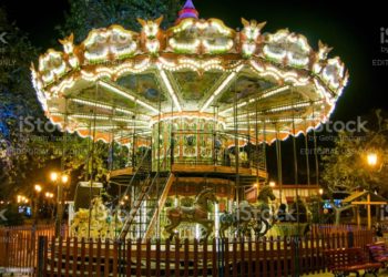 A pictiure of a big chriostmas carusel in a park,with bright lights and ,and street lights near it, which was part of a christmas decorations in the park called The christmas village,which held in a greek town thessaloniki,during the xmas season of 2019 and the early new year of 2020