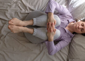 young woman working out at home, doing yoga exercise for spine on bed, lying in Knees to Chest pose, Pawanmuktasana, Gas Release Posture in the morning. Top view